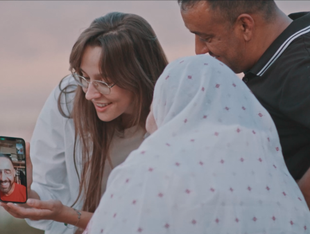 A young woman in glasses with long brown hair shows two people, a man in a black polo and a woman in a white head covering, a phone with a man in an orange shirt connected via video