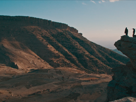 Two figures stand silhouetted on a cliff in front of a large hill and a sunset