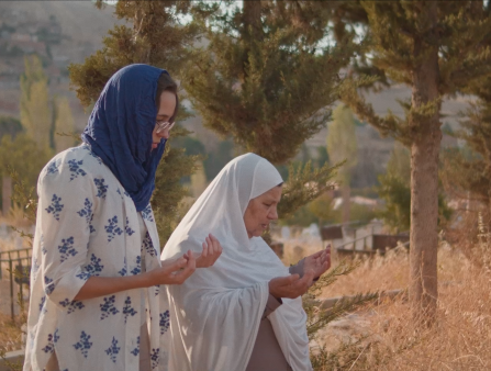 Two women in white with head coverings pray amidst tall grass