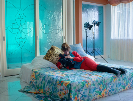 A woman in red, blue, and black lies on a mattress on the floor covered in a floral bedspread. Turquoise light shines through glass doors behind her. She holds and looks at a camera