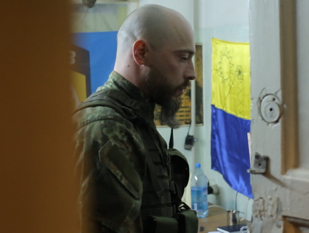A bald, bearded, uniformed soldier somberly looks down. A Ukrainian flag hangs on the wall. Shot through a doorway with wood door ajar