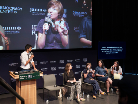 A man in a white shirt and striped tie stand behind a podium and holds a microphone to his mouth. Alongside him on stage are four women sitting in simple grey chairs and one empty grey chair. There is a large screen behind the people which displays one of the women in the chairs speaking into a microphone