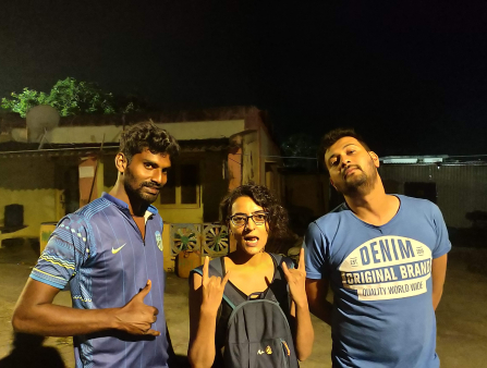 Two men stand on either side of Kaur posing for the photo at night in front of a low building. The man on the left wears a blue jersey and makes the 'hang loose' signal with his right hand. Kaur, in the middle, wears a backpack on her chest and makes the 'rock on' symbol with both hands. The man on the right, in a blue shirt which reads 'DENIM,' holds his arms behind his back.