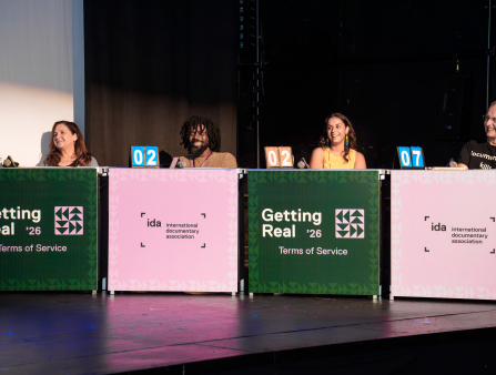 Four people sit on a stage, each behind a rectangular panel. The panels alternate between green panels which read "Getting Real '26" and pink panels which read "international documentary association." There are two women and two men, all smiling and displaying scorecards