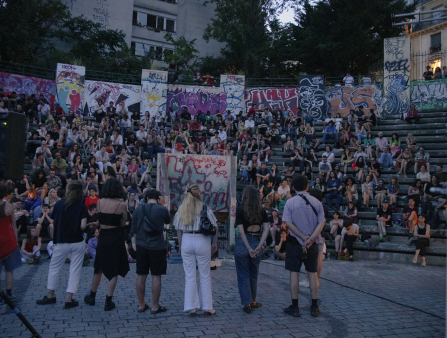 Seven people stand in a graffitied colosseum addressing a crowd which fills the seats