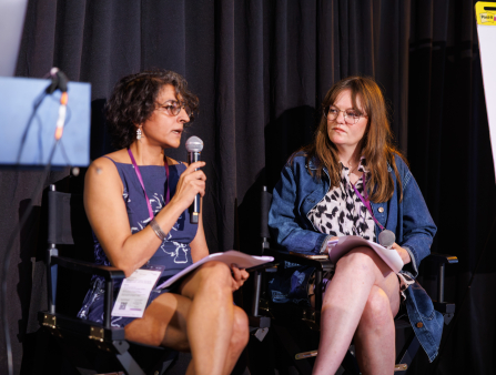 Two women sit in director's chairs next to each other in front of a black curtain, both holding microphones and paper packets. The woman on the left (Kaur) wears a floral dress, round glasses, and a lanyard around her neck. The woman on the right also wears round glasses and a purple lanyard, over a denim jacket and a collared leopard-print shirt.