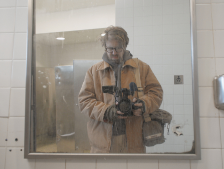 A man in an oversized light orange jacket with long brown hair and dark-rimmed glasses stands in front of a mirror holding a film camera in what looks like a public restroom