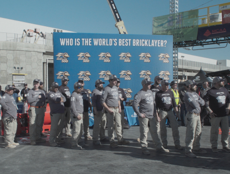 Long line of men in construction work clothes as a sign behind them reads "Who is the World's Best Brick Layer?"