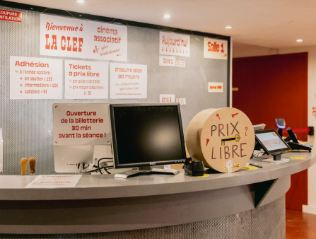 A computer, checkout kiosk, and a clock with the words "PRIX LIBRE" written on it sit on a front desk. The wall behind the desk is covered in signs welcoming guests to La Clef and providing information on ticket purchasing and screenings