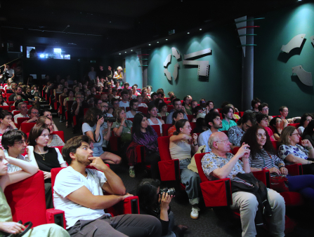 A theatre is packed with people sitting in red plush seats. The walls are blue and decorated with two large images of keys