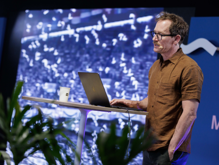 A middle-aged white man with short brown hair, a neat mustache and black-rimmed glasses stands at a podium, staring at a laptop as a screen behind him has an indistinct blurry blue-hued image