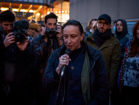 A woman in a long blue coat stands outside and speaks into a microphone. She is surrounded by people documenting her with cameras