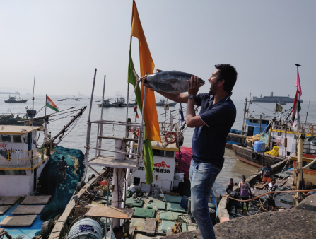 A man in jeans and a dark blue polo holds a whole tuna up to his face, appearing to sing to the fish. He stands on a concrete wall in front of a bay filled with fishing vessels of varying size.