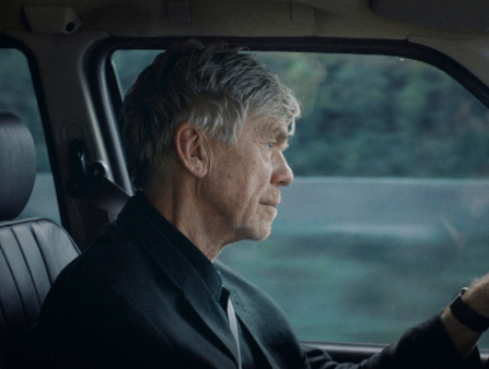 An aging white man with greyed hair, seen in profile, sits in a car wearing a suit