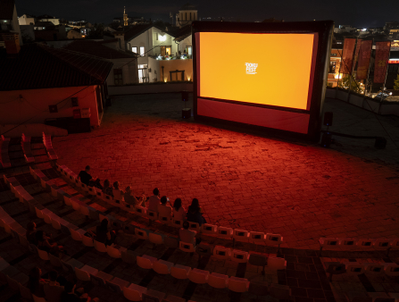 An outdoor ampitheatre watches a large orange screen which displays text reading "DOKUFEST" and bathes the space in a red glow. 