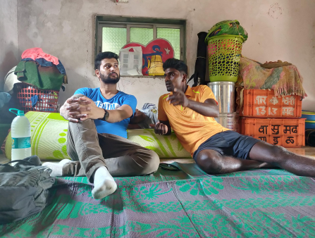 Two men sit on a floor mat in a concrete room containing various storage devices. The man on the left is bearded and wears a blue T-shirt and pants, while the man on the right is clean shaven, wearing shorts and an orange polo shirt.