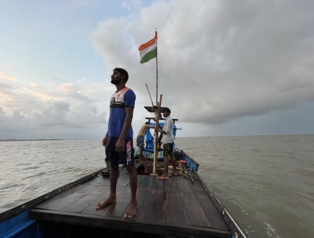 A bearded man in a jersey stands on the deck of a small boat, which holds two other men and a flagpole flying the Indian flag. Behind the boat is the open ocean and dramatic cloud cover.