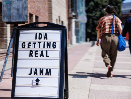 A sandwich board reads "IDA GETTING REAL JANM" with an arrow. Behind the sign, two people walk down the pavement alongside a brick building
