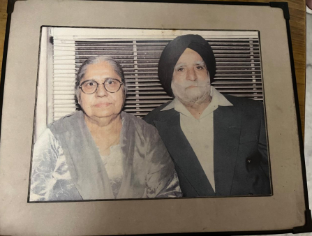 A mounted photograph of an elderly couple. On the left is a woman in a grey silk jacket and round glasses, and on the right is a man in a black suit jacket, white collared shirt, and head scarf.