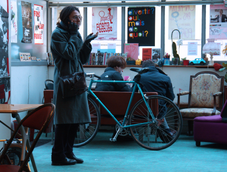A man in a trench coat stands in a room filled with natural light, speaking on the phone and gestures with gloves hands. Behind him, a bicycle rests against a couch on which two figures sit hunched over. The walls are lined with books and posters