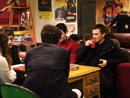 Five people dressed warmly sit around a yellow wooden table. Behind them are a leather couch and walls lined with film posters