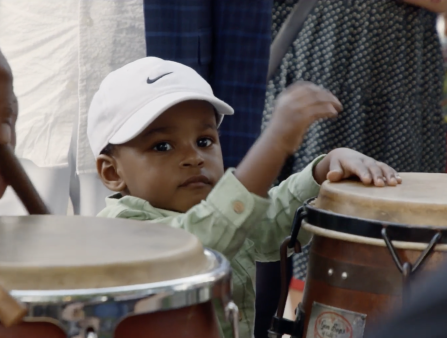 A little boy in a white Nike cap and a green, plaid, button-down shirt plays a drum almost as tall as he is with his hands. In the foreground, a pair of adult hands play a similar drum using short sticks