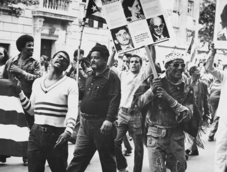 Black and white photo of a  large crowd of men marching down a city street,  holding posters of people's faces and the Puerto Rican flag.