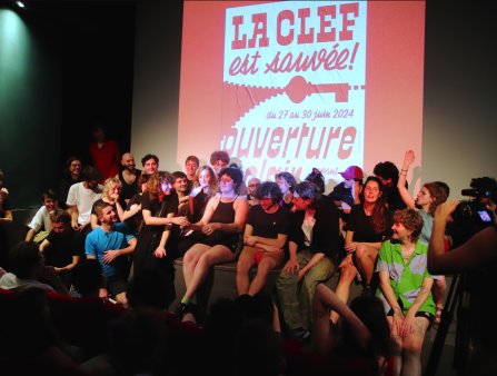 A small stage is packed with joyous people in colorful clothing. A large screen behind them displays a poster which reads "LA CLEF est sauvée!"
