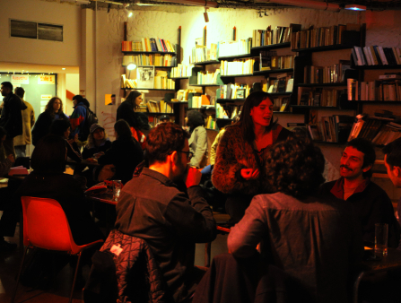 A group of nicely dressed people sit around a table illuminated in red light. More people stand and sit around the perimeter of the room, which is lit in yellow and contains walls lined with books