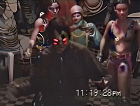 Low resolution image in which a person in a dark jacket with four glowing, red eyes stands in front of three other uncanny figures, each falling somewhere in between mannequin and human and dressed in eccentric, colorful costumes. The room they are in has a black and white checkered floor. A timestamp at the bottom right of the screen reads 11:19:28PM