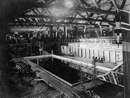 Black and white image of a large, empty indoor swimming pool, with wooden beams exposed and flags hanging from the ceiling