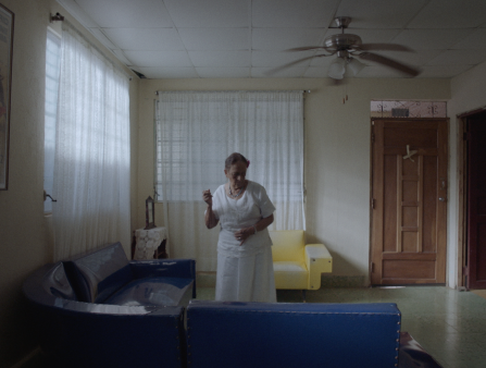 An elderly woman in a white dress dances in a living room which contains a blue couch, a yellow couch, and large windows covered by white curtains