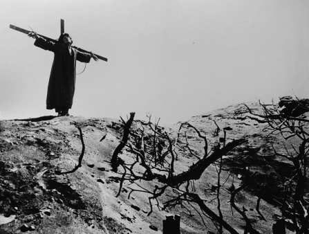 Black and white image of a figure in a dark robe on a crucifix atop a barren hill covered in sparse, dead vegetation