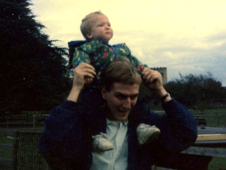 A man smiles and holds the hands of a small child who sits on his shoulder. The man wears a dark blue jacket over a light blue buton-down shirt, and the young child wears a green jumpsuit with colored shapes. The pair are on a farm with horses in a fenced enclosure visible behind them, as well as large, dark conifers and a cloudy sky at dusk