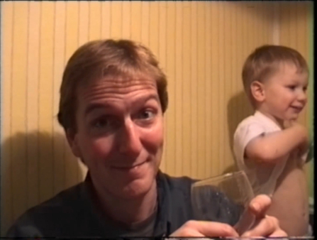 A man with short brown hair smiles awkwardly and holds up a glass. To his left is a smiling young boy lifting up his shirt. The walls of the room are wood panelled
