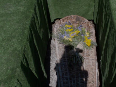 A shadow of a woman holding a camera is visible on a woven wooden coffin decorated with yellow and blue flowers. The coffin is underground, and the ground , both around and above the coffin, appears to be green turf