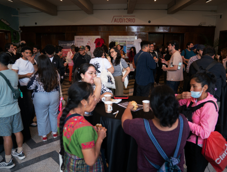 Groups of colorfully dressed people stand around an auditorium conversing. In the foreground, women with disposable coffee cups gather around a round table covered in a black tablecloth