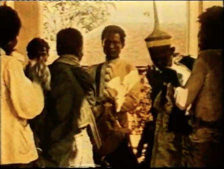 Orange image of three men with their backs to the camera facing three men who face the camera. Some are dressed in robes, while the man in the center wears a military jacket. A man on the right wears a tall, skinny pointed hat 