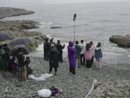 A film crew of about 20 people in raincoats, some of whom hold umbrellas, stand on a rocky shore, capturing image of a woman in a pink dress, who sits on a rock surrounded by waves
