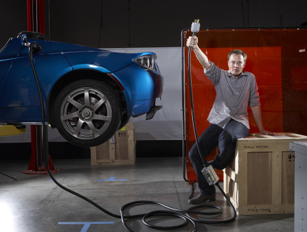 Musk wears a grey button down shirt and sits on a wooden crate in a garage.  He holds a plug over his head triumphantly which is connected to a blue Tesla suspended in midair.