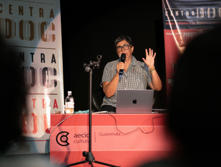 A woman with short hair, glasses and a short-sleeved, striped, button-down shirt sits behind a red podium with an open macbook and water bottle on it and speaks into a microphone. Two banners behind her read "CENTRADOC"