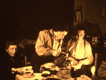 Low resolution yellowed footage of three people dressed nicely sitting around a dinner table. One is a woman who stands and pours liquid into the cup of a seated man. There is a tablecloth over the table and fancy dishes