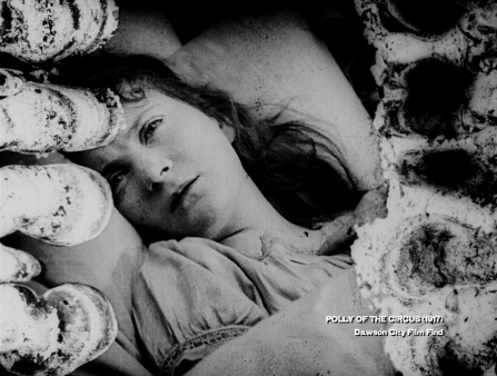 Black and white image of the face and shoulders of a woman laying in bed. Damage to the film has produced unusual circular shapes on the left and righthand sides of the image. A caption in the lower righthand corner reads "POLLY OF THE CIRCUS (1917) Dawson City Film Find"