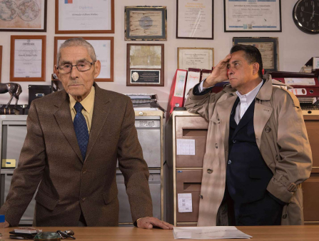 An elderly man in a brown suit and a middle-aged man in trench coat are behind an office desk