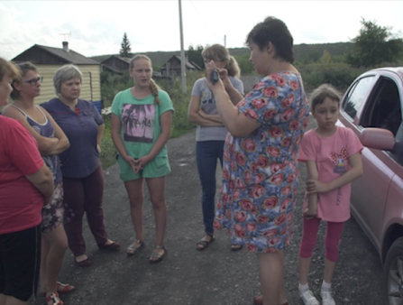 Homemaker-turned-journalist Natalia Zubkova in a blue and pink flowered dress, interviews five women with her cell phone in a rural village as her young daughter, in pink shirt and capri pants, waits nearby.