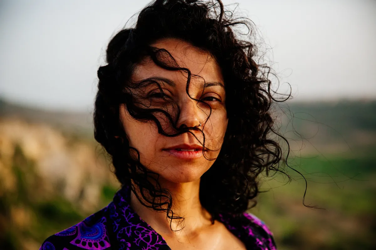 A woman with brown skin stares at the camera. Her dark brown curly hair drapes over her face, and a black and purple shawl is wrapped around her shoulders.
