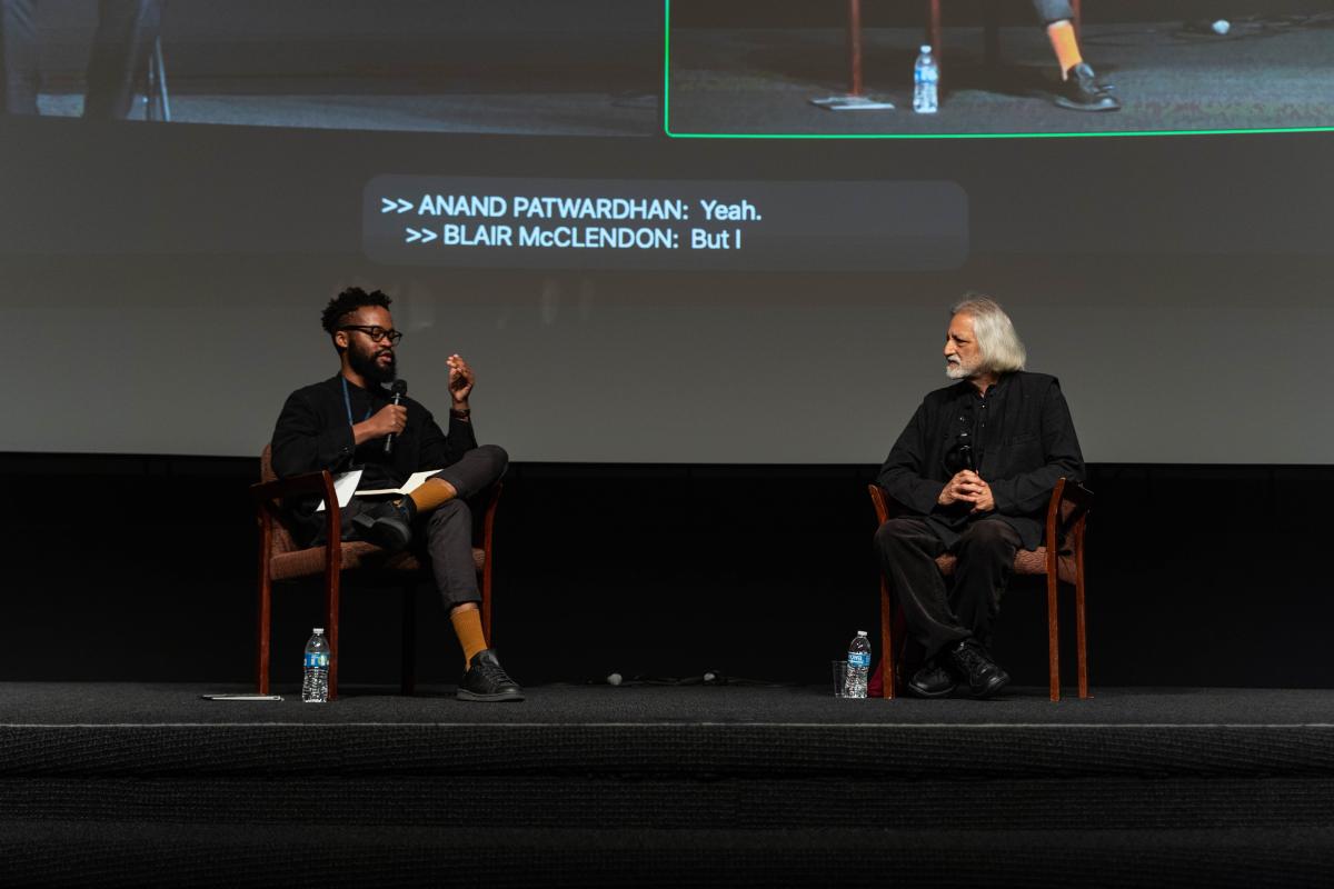 two people sitting in chairs on a stage. one person, dark brown skin, black glasses, black hair, dressed in all black, the other is fair skin complexion, with white hair, dress in all black