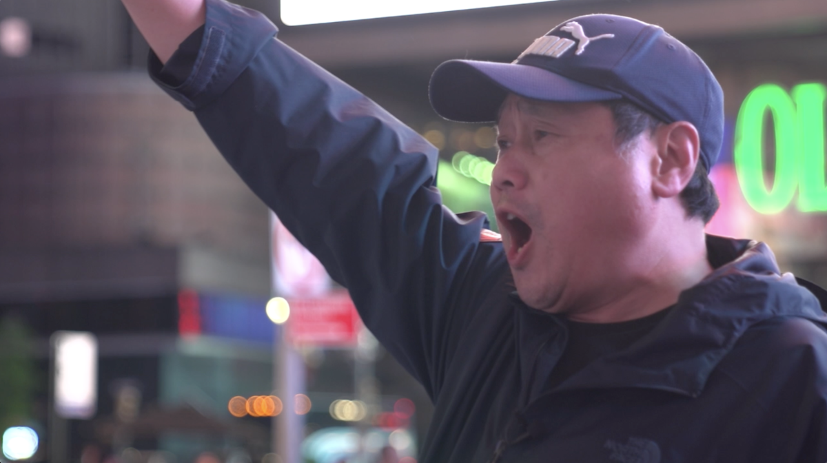 A protesting man wearing a dark blue sweater and match baseball cap thrusting his right arm during a protest in Times Square NYC.