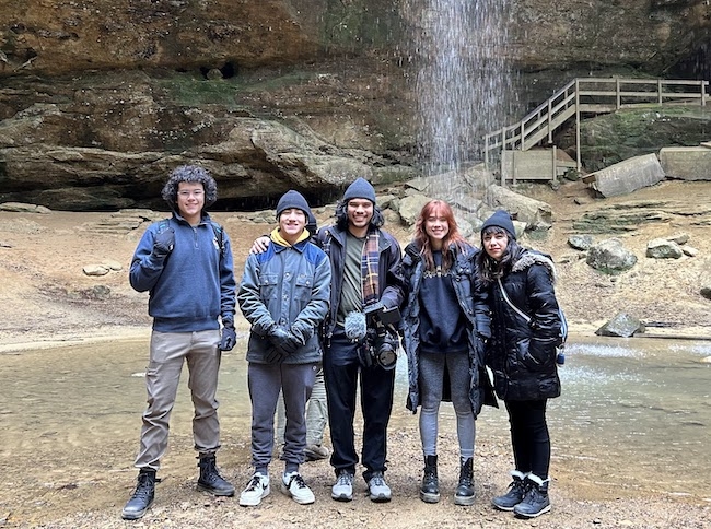 Five donor-conceived siblings wearing winter clothes stand in front of a waterfall. 
