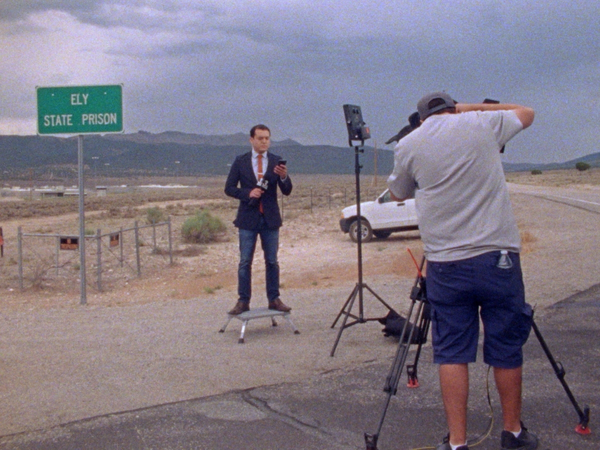 A news anchor stands in the middle of the desert, in front of a sign that says, “ELY STATE PRISON”. A production light shines on them, and a camera person with a tripod films them. 